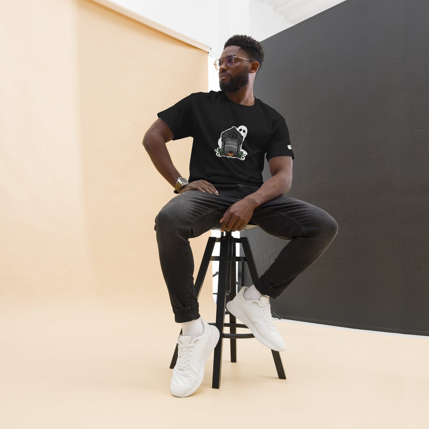 Product: Black t-shirt with a centered design of a gravestone that reads "here lies transphobia" with a ghost and fall foliage surrounding the gravestone. This is image is the product on a straight-sized male model.