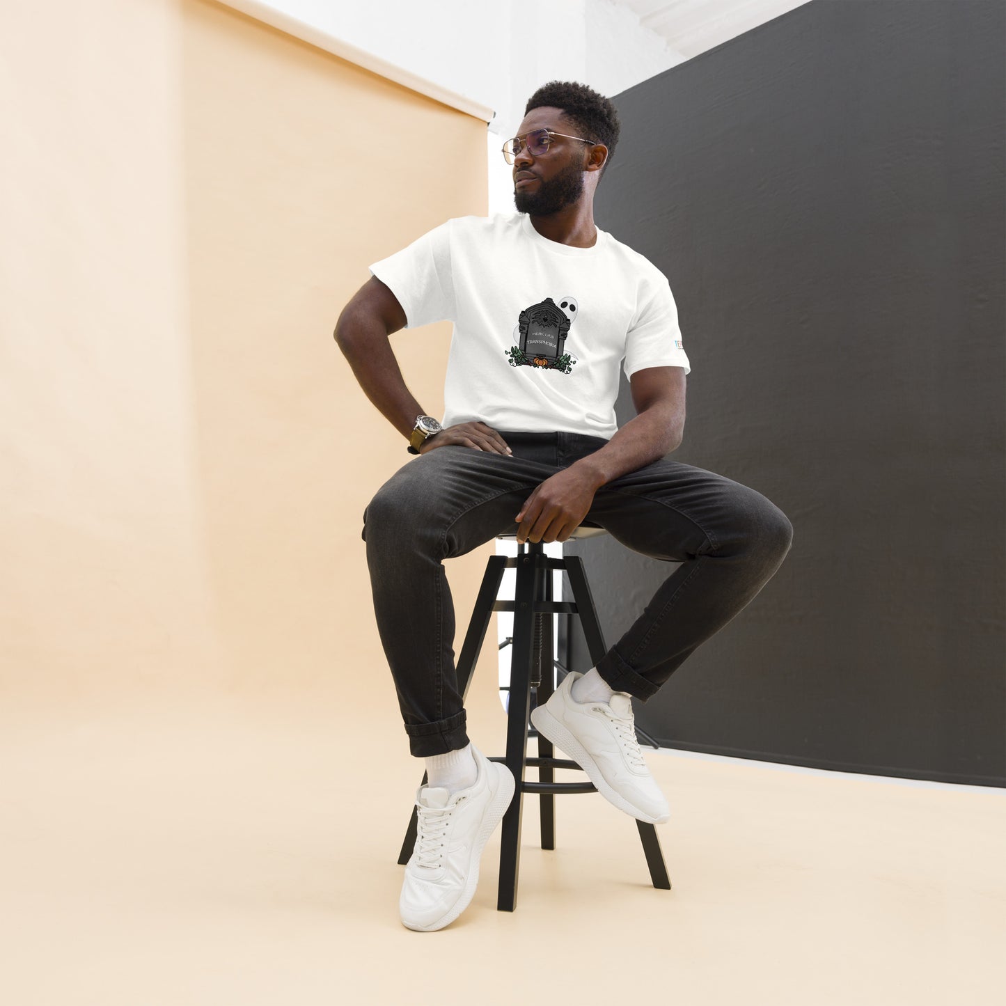 Product: White t-shirt with a centered design of a gravestone that reads "here lies transphobia" with a ghost and fall foliage surrounding the gravestone. This is image is the product on a straight-sized male model.