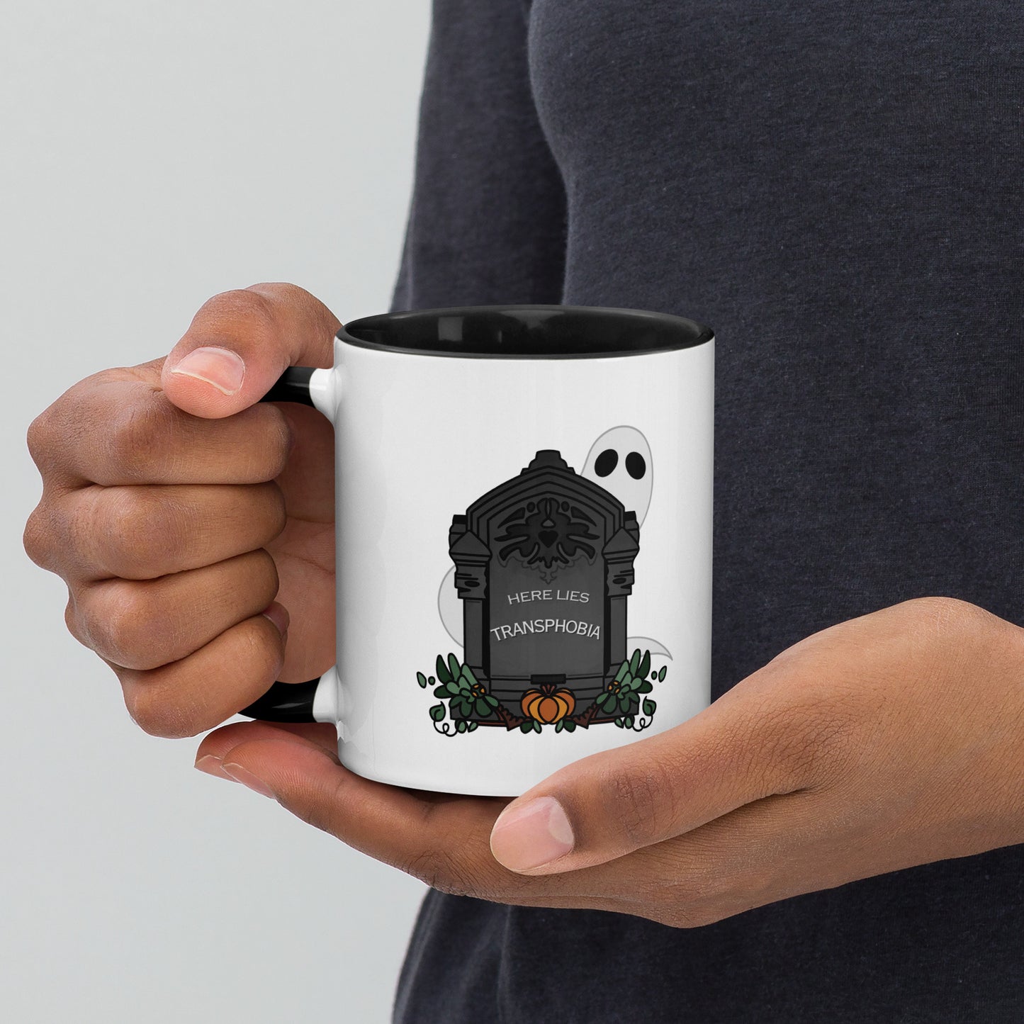 Front view of a white mug with an image gravestone and ghost, with the text "here lies transphobia" in white on the gravestone. The handle and inside of the mug are black.