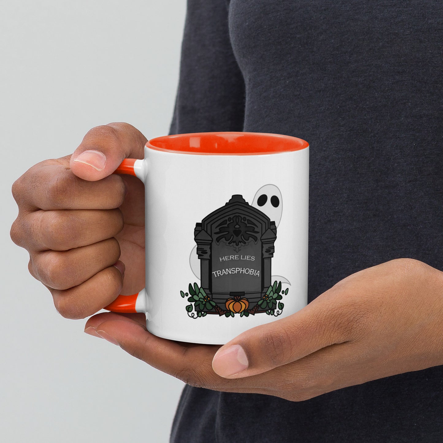 Front view of a white mug with an image gravestone and ghost, with the text "here lies transphobia" in white on the gravestone. The handle and inside of the mug are orange.