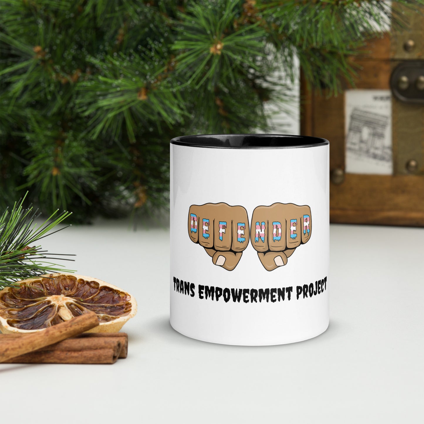 Front view of a white mug with an image of fists with "defender" written across the knuckles in blue, pink, and white. and "TRANS EMPOWERMENT PROJECT" in black lettering below the image. the inside of the mug is black.