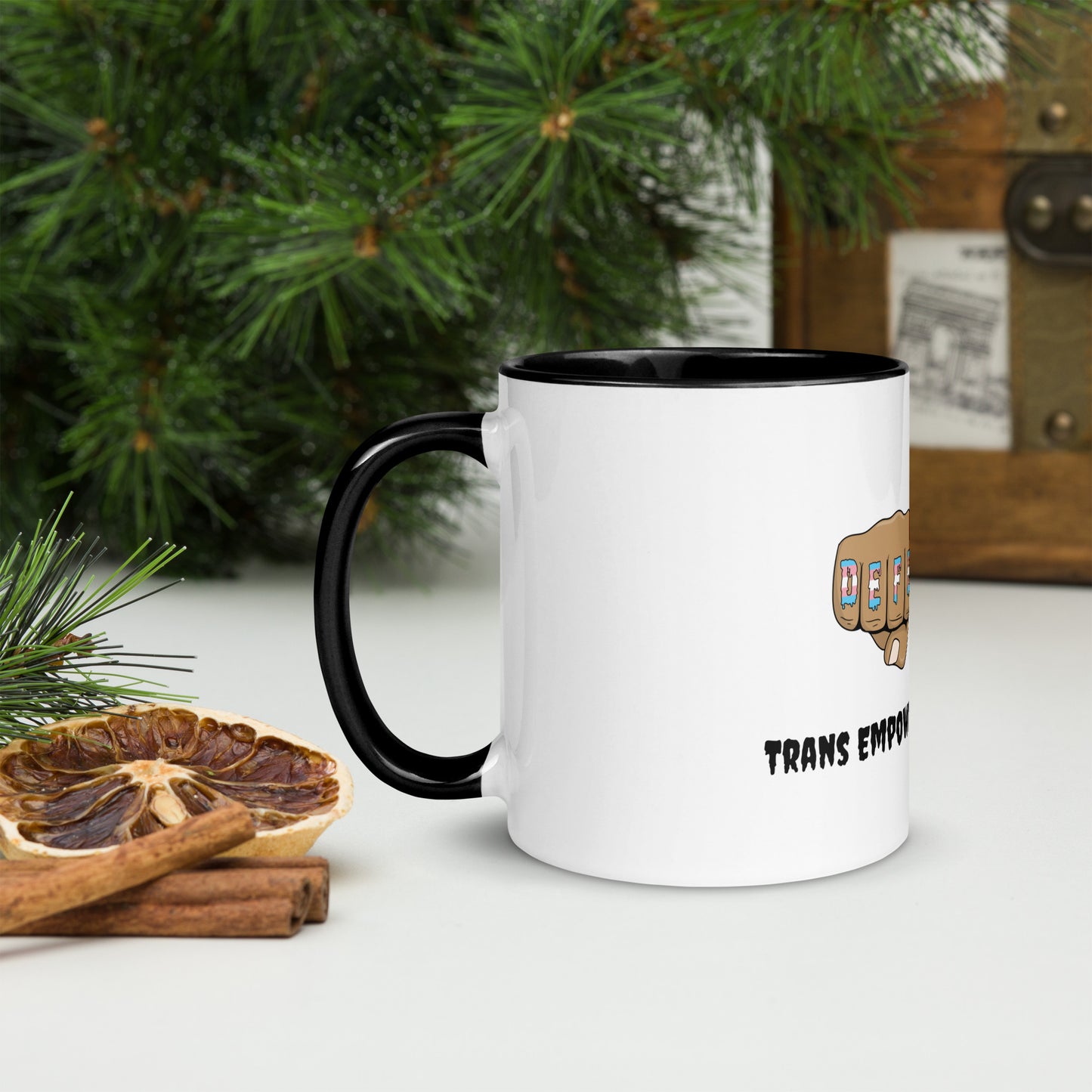 Side view of a white mug with an image of fists with "defender" written across the knuckles in blue, pink, and white. and "TRANS EMPOWERMENT PROJECT" in black lettering below the image. The handle and inside of the mug are black.