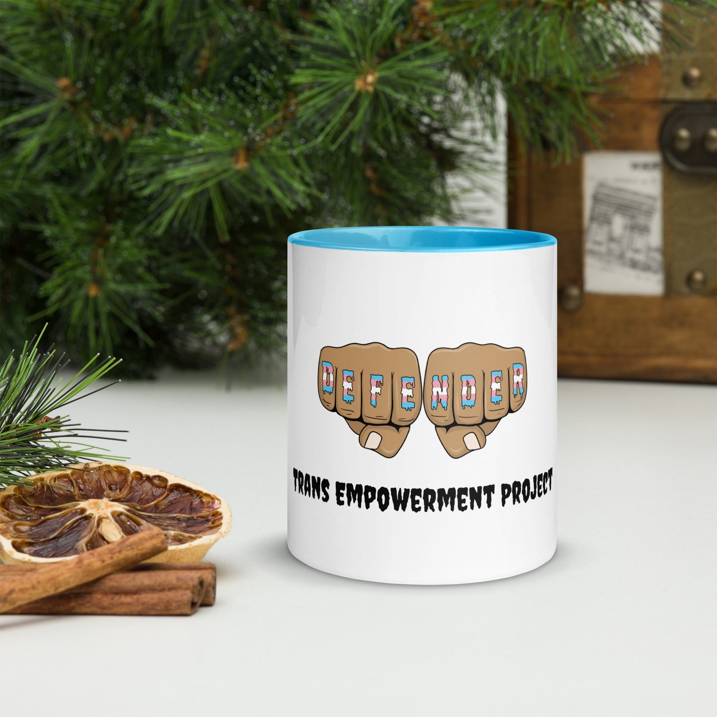 Front view of a white mug with an image of fists with "defender" written across the knuckles in blue, pink, and white. and "TRANS EMPOWERMENT PROJECT" in black lettering below the image. The handle and inside of the mug are light blue.
