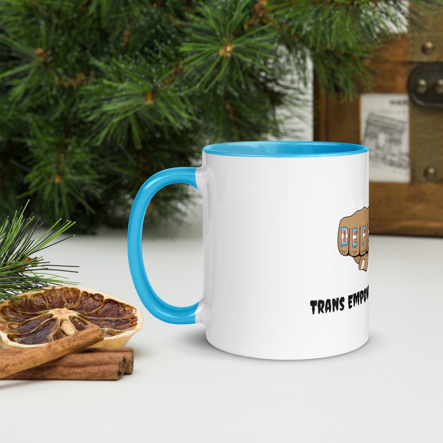 Side view of a white mug with an image of fists with "defender" written across the knuckles in blue, pink, and white. and "TRANS EMPOWERMENT PROJECT" in black lettering below the image. The handle and inside of the mug are light blue.