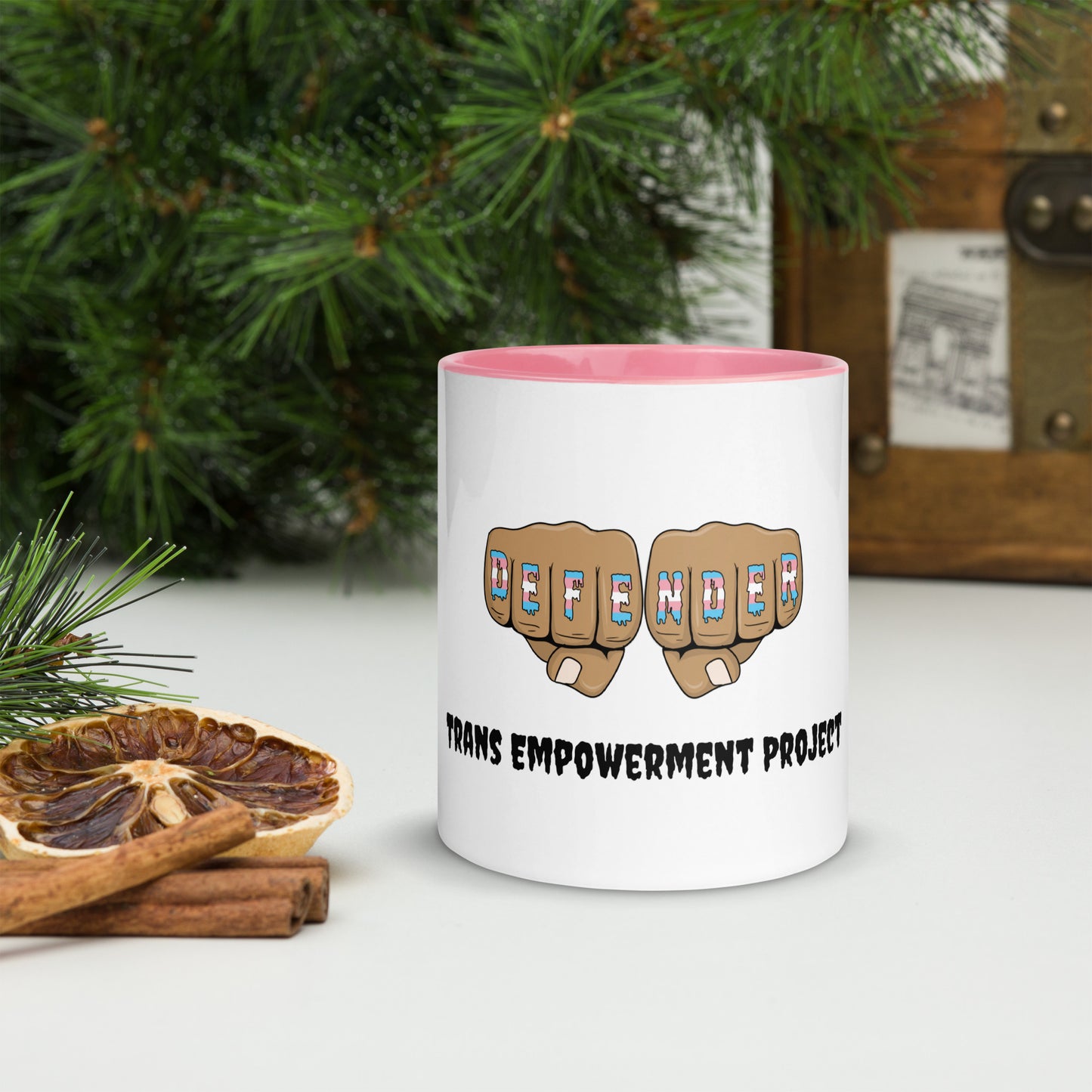 Front view of a white mug with an image of fists with "defender" written across the knuckles in blue, pink, and white. and "TRANS EMPOWERMENT PROJECT" in black lettering below the image. The handle and inside of the mug are light pink.