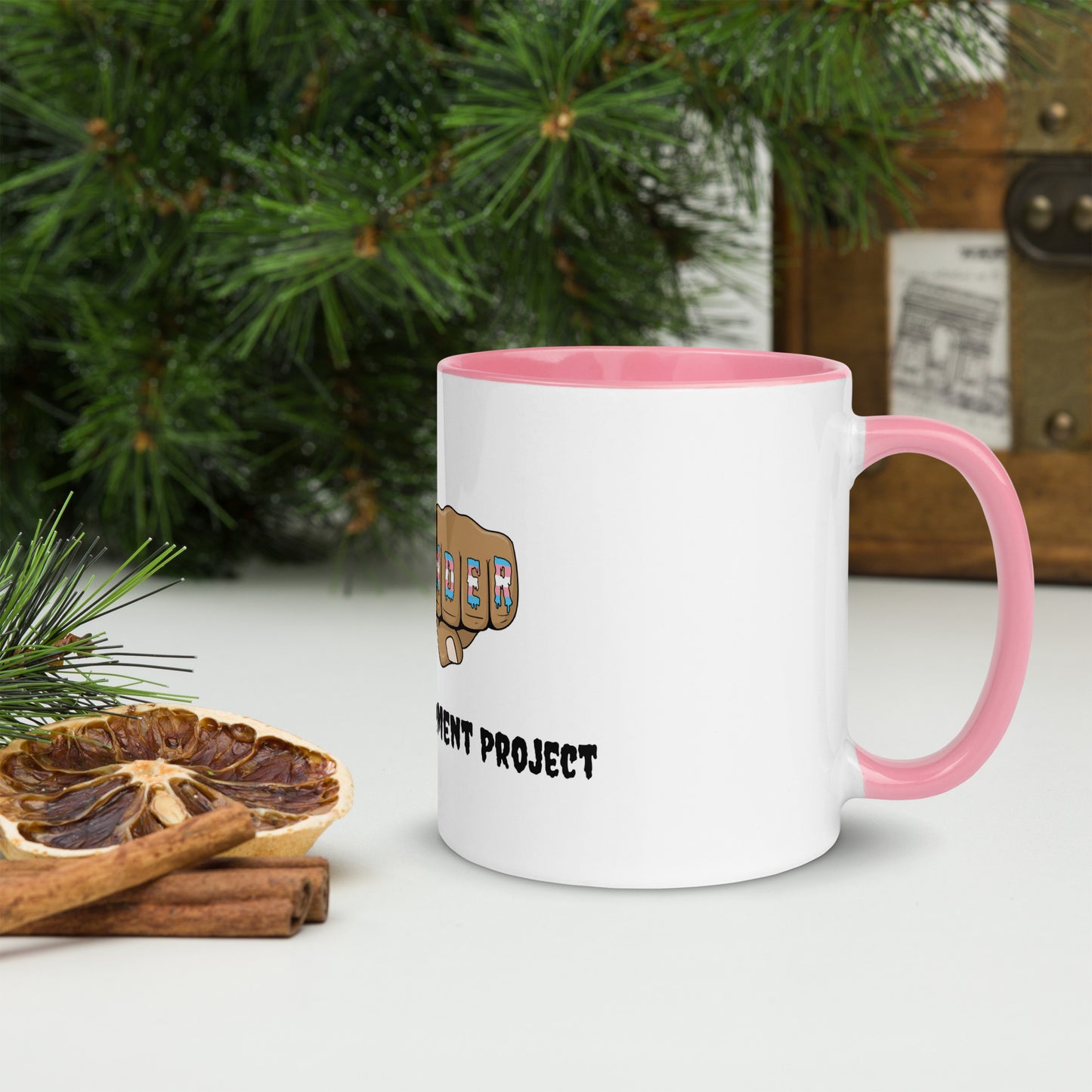 Side view of a white mug with an image of fists with "defender" written across the knuckles in blue, pink, and white. and "TRANS EMPOWERMENT PROJECT" in black lettering below the image. The handle and inside of the mug are light pink.