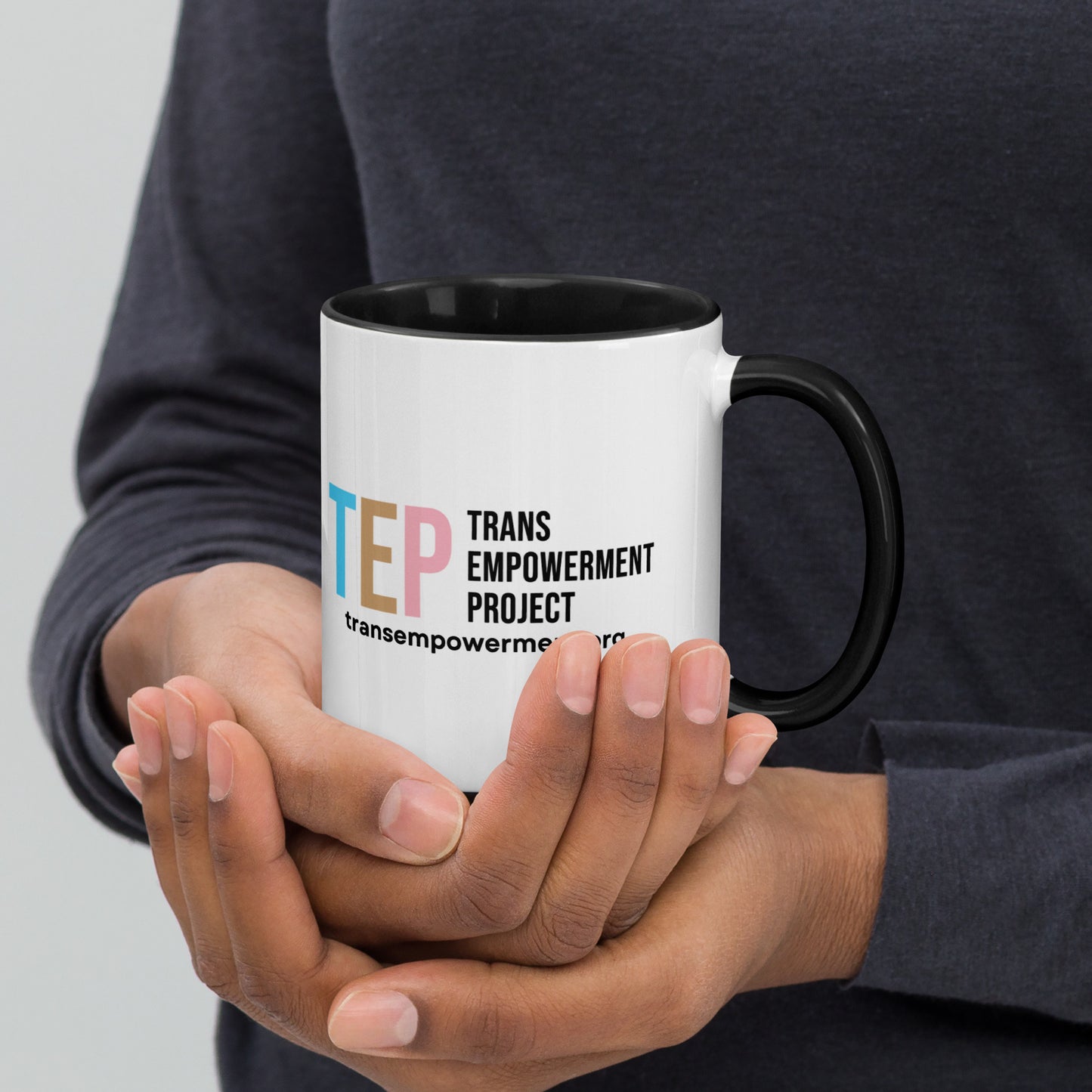 Reverse side view of a white mug with the Trans Empowerment Logo and the text "transempowerment.org" underneath. The handle and inside of the mug are black.