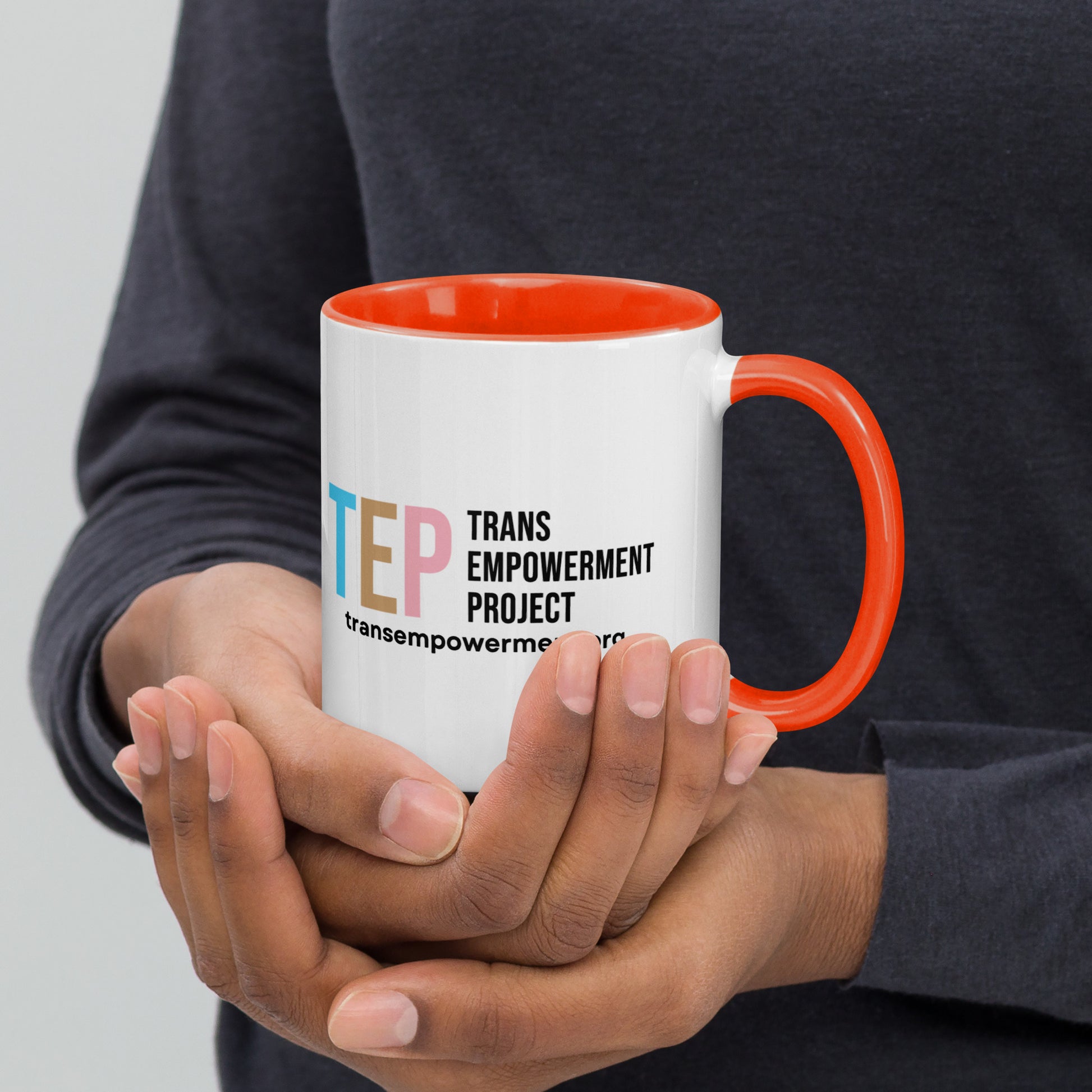 Reverse side view of a white mug with the Trans Empowerment Logo and the text "transempowerment.org" underneath. The handle and inside of the mug are orange.