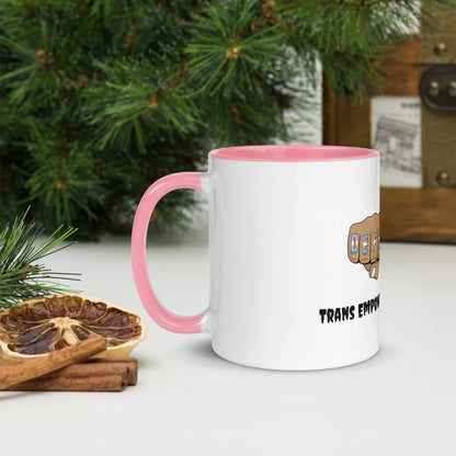 Side view of a white mug with an image of fists with "defender" written across the knuckles in blue, pink, and white. and "TRANS EMPOWERMENT PROJECT" in black lettering below the image. The handle and inside of the mug are light pink.